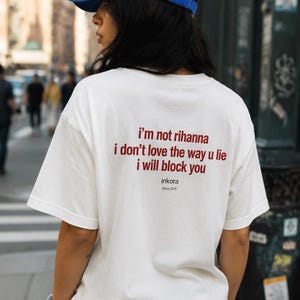 Puede incluir: Camiseta blanca con el texto rojo "i'm not rihanna i don't love the way u lie i will block you". La camiseta también tiene el texto "inkora Since 2025". Se lleva una gorra de béisbol azul con "BALANCED".