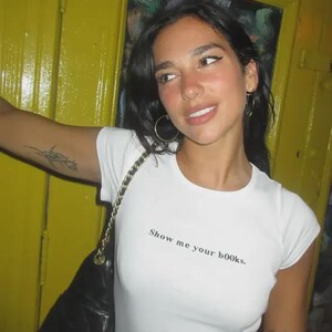 May include: A white short-sleeved t-shirt featuring the black text "Show me your b00ks." printed on the chest. The shirt has a crew neckline and a fitted silhouette. A black quilted shoulder bag with a gold chain strap is worn over the shoulder.