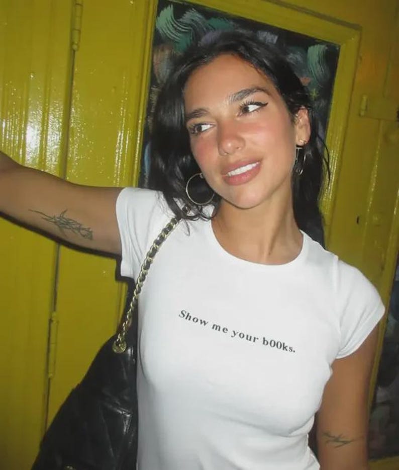 May include: A white short-sleeved t-shirt featuring the black text "Show me your b00ks." printed on the chest. The shirt has a crew neckline and a fitted silhouette. A black quilted shoulder bag with a gold chain strap is worn over the shoulder.