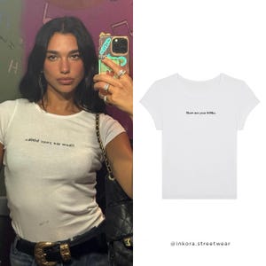 May include: A white short-sleeved t-shirt featuring the black text "Show me your b00ks." printed on the chest. The garment has a round neckline and a fitted silhouette, showcasing a simple design with clear, legible lettering across the front.