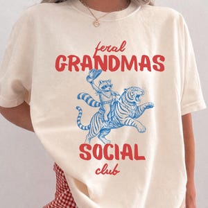 May include: Ivory-colored t-shirt with the text "feral GRANDMAS SOCIAL club" in red. A blue illustration depicts a raccoon riding a tiger. The shirt is a relaxed fit.