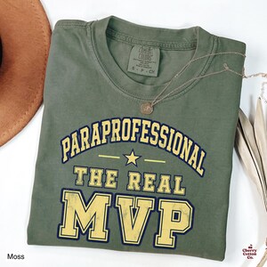 May include: A moss green t-shirt featuring the text PARAPROFESSIONAL THE REAL MVP in a collegiate font with a yellow star graphic. The shirt has a crew neckline and a Comfort Colors brand label on the inside collar indicating a size small.