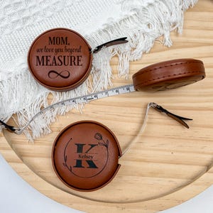 Può includere: Metro a nastro in similpelle marrone con la scritta "MOM, we love you beyond MEASURE" e un simbolo di infinito. Un altro nastro ha il nome "Kelsey" con un design floreale. Il nastro misura fino a 150 cm.