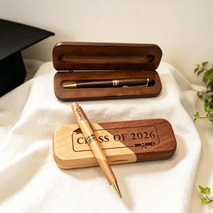 Può includere: Set di penna e astuccio in legno a tema laurea. Una penna è esposta all'interno di un astuccio in legno scuro, e un'altra penna poggia su un astuccio bicolore con inciso "Class of 2026". Un cappello di laurea con nappina dorata è sullo sfondo.