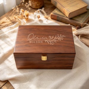 May include: A rectangular dark brown wooden keepsake box with a gold-colored metal latch. The lid features the engraved name Olivia Wilson in a script and serif font, accompanied by a delicate line-art floral branch illustration.
