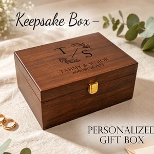 Könnte beinhalten: Eine rechteckige Holz-Andenkenbox mit goldenem Verschluss. Die Box ist mit Initialen, Namen und einem Datum graviert. Der Text "Keepsake Box" und "Personalized Gift Box" sind ebenfalls sichtbar.