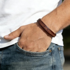 May include: A brown leather bracelet inscribed with the words "FOUND MY TREASURE". The bracelet is worn on a wrist. The person is wearing a white t-shirt and blue jeans.
