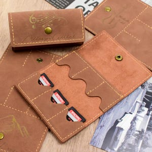 Puede incluir: Portatarjetas SD de cuero marrón con cierre de botón a presión. El soporte tiene tres ranuras para tarjetas y está cosido con un hilo en contraste. La parte superior del soporte tiene un diseño de notas musicales y el nombre "Allison Culler".