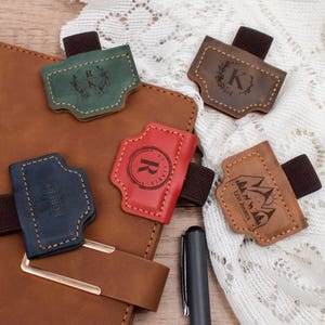 May include: A collection of leather journal pen holders in various colors, including green, brown, red, and navy blue. Each holder features a unique design, such as initials, a mountain scene, or a name, and is attached to an elastic band.