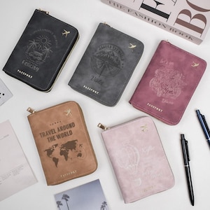 May include: Five passport holders in various colors: dark gray, gray, mauve, tan, and light pink. Each has a gold zipper and a small airplane emblem. The covers feature travel-themed designs and text, including "Felicity," "Violet," and "Travel Around The World."
