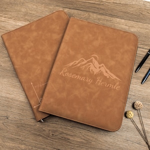 May include: Two brown leather document holders with a mountain and sun design and the name "Rosemary Hermle" engraved on one. The other holder has a compass design. Pens and decorative items are on the wooden surface.
