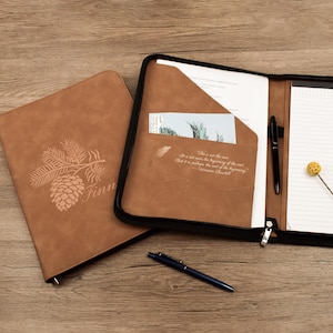 Può includere: Due cartelle in pelle marrone, una chiusa e una aperta. La cartella chiusa presenta un design a pigna e ramo con il nome "Finn" in rilievo. La cartella aperta ha una cerniera nera, una tasca con una carta e una penna. Una penna blu è appoggiata sulla superficie di legno.