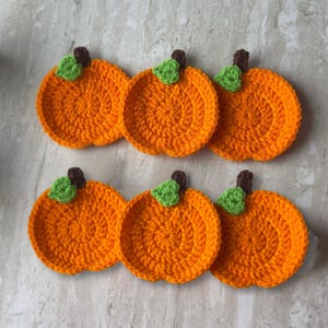 Könnte beinhalten: Ein Set aus sechs runden, orangefarbenen Häkeluntersetzern, die jeweils mit einem kleinen grünen Blatt und einem kurzen braunen Stiel versehen sind. Die Untersetzer sind in zwei Reihen zu je drei Stück angeordnet und zeigen ein strukturiertes Garnmuster.