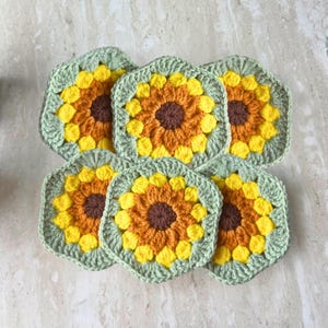 May include: Five square crochet coasters featuring a sunflower motif. Each coaster has a dark brown centre, a ring of orange petals, a ring of bright yellow petals, and a sage green border. The pieces are arranged in a cluster on a surface.