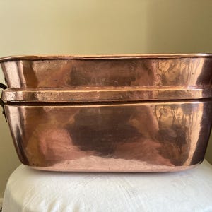 May include: Two antique copper tubs, one stacked on top of the other. The tubs have a rectangular shape with rounded edges and dark metal handles. The copper has a warm, reddish-brown hue with visible patina.