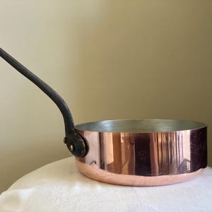 May include: A copper saucepan with a dark, curved handle. The pan has a shiny, reflective surface and a silver interior. The handle is attached with rivets. This cookware is suitable for cooking.