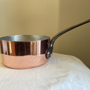 Könnte beinhalten: Ein Kupfertopf mit einem langen, dunklen Griff. Der Topf hat eine glänzende Kupferaußenseite und eine silberne Innenseite. Der Griff ist mit Kupfernieten befestigt. Die Pfanne steht auf einer weißen Oberfläche.