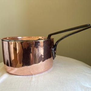 May include: A copper saucepan with a dark metal handle. The pot has a shiny, reflective surface and is resting on a white surface. The handle extends out from the side of the pot.