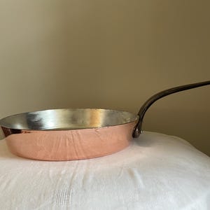Könnte beinhalten: Eine Kupferpfanne mit einem langen, dunklen Griff. Die Pfanne hat einen glänzenden, silbernen Innenraum und eine Kupferaußenseite. Die Pfanne steht auf einer weißen Oberfläche. Die Pfanne wird wahrscheinlich zum Kochen verwendet.