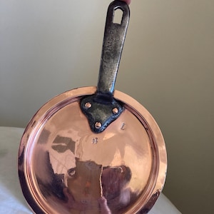 Op de afbeelding: Een koperkleurige, ronde koekenpan met een donker metalen handvat. Het handvat is aan de pan geklonken en heeft de tekst "C. ENAG. P." erop gestempeld. Het nummer "14" is op het oppervlak van de pan gestempeld.