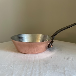 Op de afbeelding: Een koperen steelpan met een lange, donkere handgreep. De binnenkant van de pan is zilverkleurig en de buitenkant is gepolijst koper. De pan staat op een wit oppervlak.