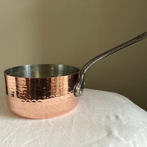 Pode incluir: Uma panela de cobre com acabamento martelado e uma longa alça de metal rebitada. O interior é prateado. A panela está sobre uma superfície branca. Esta panela é ideal para cozinhar e servir.