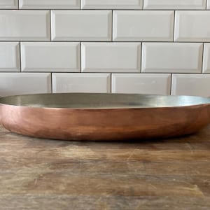 Pode incluir: Uma frigideira de cobre larga e rasa com um revestimento interior prateado metálico e duas pegas curvas de latão fixadas nos lados opostos. O exterior apresenta um acabamento em cobre polido, enquanto o interior mostra uma superfície metálica lisa.
