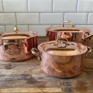 Peut inclure: Un ensemble de trois casseroles en cuivre poli avec couvercles assortis et poignées en laiton. Les casseroles sont rondes avec une finition lisse et réfléchissante. Chaque couvercle possède une poignée centrale en laiton.