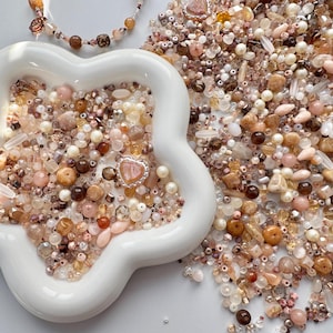 May include: A white, star-shaped dish filled with an assortment of beads in pink, brown, and cream hues. A beaded bracelet and necklace are also visible. The beads are of various sizes and shapes, including round, oval, and irregular forms, creating a textured and colourful display.