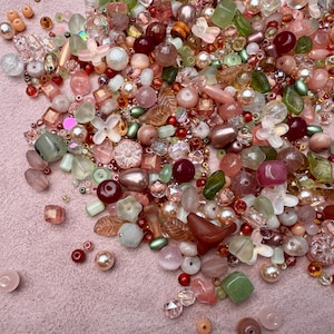 May include: A large collection of loose craft beads in various shades of pink, green, red, and clear. The assortment includes round, faceted, leaf-shaped, and flower-shaped beads made of glass, plastic, and stone with diverse textures and finishes.