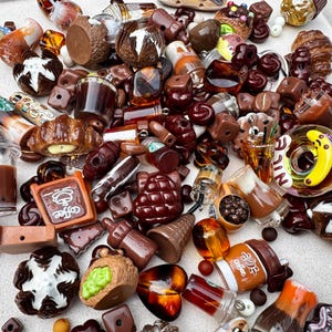May include: A collection of miniature food and beverage charms including coffee cups, croissants, donuts, and chocolate pieces. The items feature brown, amber, and white tones with detailed textures, some displaying the word coffee or a banana graphic.