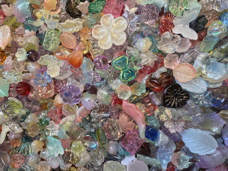 May include: A large collection of assorted acrylic and glass beads in various shapes, including flowers, butterflies, hearts, and leaves. The beads feature a mix of translucent, opaque, iridescent, and solid colors like pink, blue, green, and clear.