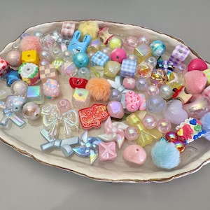 Peut inclure: Un assortiment de perles et de breloques colorées de différentes formes et tailles, dont des cubes, des cœurs, des nœuds et des pompons. Les perles sont dans des tons de rose, de bleu, de jaune et irisé, disposées sur un plat décoratif de forme ovale.