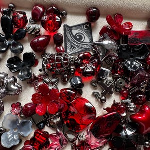 May include: A collection of assorted beads and charms in shades of red, black, and metallic silver. The assortment includes flower-shaped beads, star-shaped charms, spherical beads, and a square pendant featuring a graphic illustration of an eye.