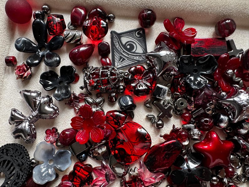 May include: A collection of assorted beads and charms in shades of red, black, and metallic silver. The assortment includes flower-shaped beads, star-shaped charms, spherical beads, and a square pendant featuring a graphic illustration of an eye.
