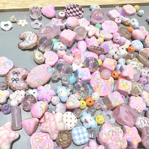 Pode incluir: Uma cole&ccedil;&atilde;o de contas e berloques em cores pastel em v&aacute;rias formas, incluindo estrelas, flores e donuts. As contas s&atilde;o em tons de rosa, roxo e azul, algumas com padr&otilde;es xadrez e detalhes brilhantes. Existe uma variedade de tamanhos e formas.