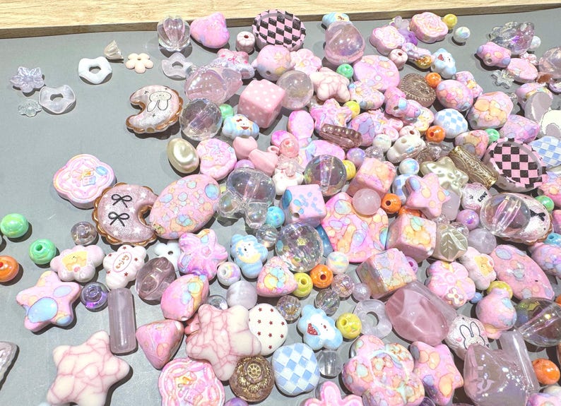 Pode incluir: Uma cole&ccedil;&atilde;o de contas e berloques em cores pastel em v&aacute;rias formas, incluindo estrelas, flores e donuts. As contas s&atilde;o em tons de rosa, roxo e azul, algumas com padr&otilde;es xadrez e detalhes brilhantes. Existe uma variedade de tamanhos e formas.