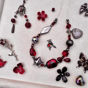 May include: A beaded necklace featuring red and silver-toned charms, including a bird, a cross, and a heart, surrounded by loose components like a rose-shaped pendant, a butterfly with skull patterns, flower beads, and a small birdcage charm.
