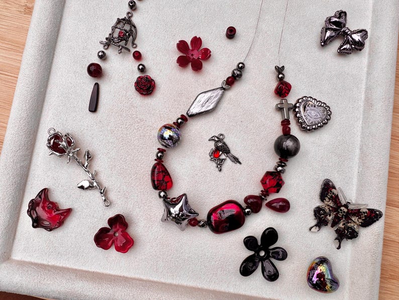 May include: A beaded necklace featuring red and silver-toned charms, including a bird, a cross, and a heart, surrounded by loose components like a rose-shaped pendant, a butterfly with skull patterns, flower beads, and a small birdcage charm.