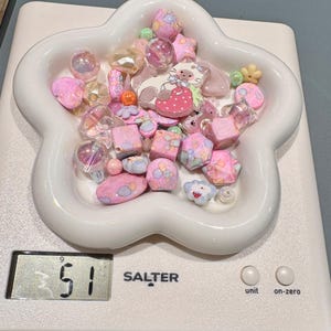 Pode incluir: Uma cole&ccedil;&atilde;o de contas rosa e pastel numa tigela branca em forma de flor, sobre uma balan&ccedil;a digital. As contas incluem v&aacute;rias formas como estrelas, cubos e esferas, com um berloque com tema de gato. A balan&ccedil;a mostra um peso de 51.