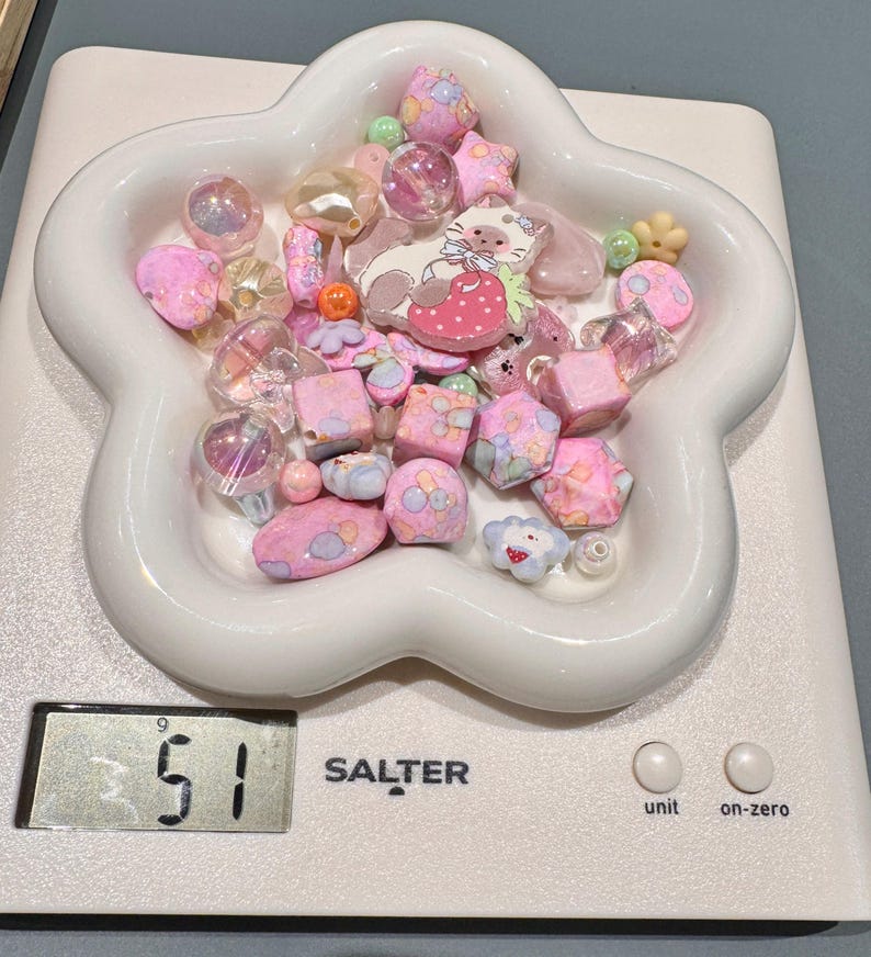 Pode incluir: Uma cole&ccedil;&atilde;o de contas rosa e pastel numa tigela branca em forma de flor, sobre uma balan&ccedil;a digital. As contas incluem v&aacute;rias formas como estrelas, cubos e esferas, com um berloque com tema de gato. A balan&ccedil;a mostra um peso de 51.