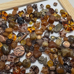May include: A selection of brown and amber-toned beads and decorative items. The collection features beads in diverse shapes, sizes, and textures, including round, square, and star-shaped. Some beads display detailed patterns and metallic highlights.