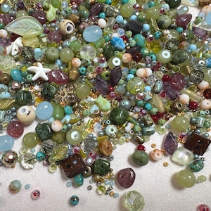 May include: A collection of assorted loose beads in various shapes, sizes, and textures, featuring shades of green, blue, brown, and clear glass. The mix includes round, faceted, leaf-shaped, and star-shaped beads, along with small metallic spacers.