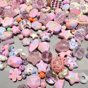 Pode incluir: Uma cole&ccedil;&atilde;o de contas e berloques rosa e iridescentes em v&aacute;rias formas, incluindo estrelas, flores e cora&ccedil;&otilde;es. Alguns berloques t&ecirc;m detalhes e padr&otilde;es pintados. As contas est&atilde;o espalhadas sobre uma superf&iacute;cie cinza.