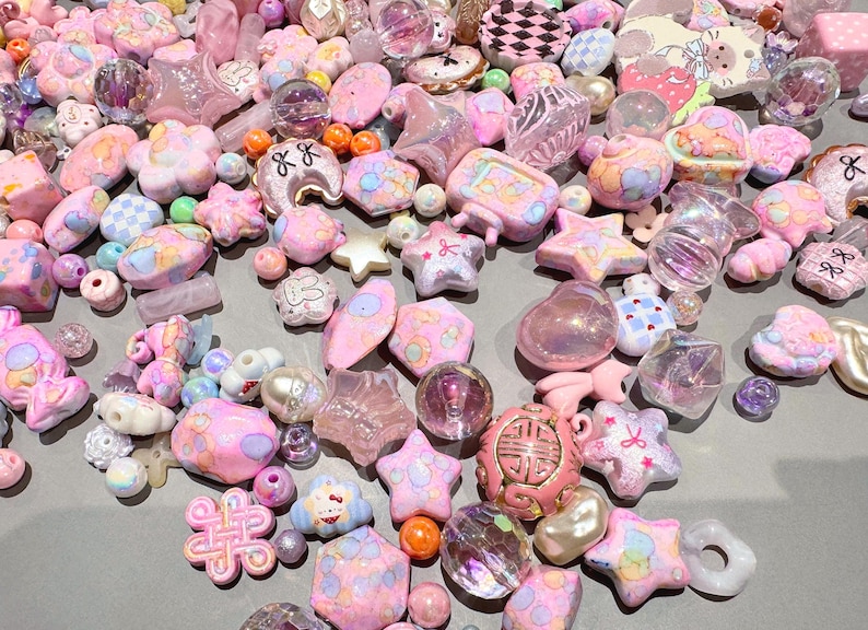 Pode incluir: Uma cole&ccedil;&atilde;o de contas e berloques rosa e iridescentes em v&aacute;rias formas, incluindo estrelas, flores e cora&ccedil;&otilde;es. Alguns berloques t&ecirc;m detalhes e padr&otilde;es pintados. As contas est&atilde;o espalhadas sobre uma superf&iacute;cie cinza.