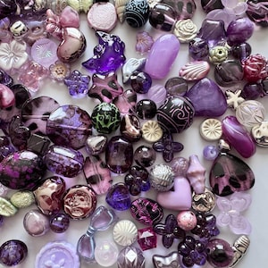 Puede incluir: Una colección de diversas cuentas moradas e iridiscentes, incluyendo diseños redondos, ovalados y en forma de flor. Las cuentas varían en tamaño y textura, algunas con patrones intrincados y otras lisas y pulidas. La paleta de colores va del lavanda claro al violeta oscuro.