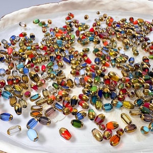 May include: A selection of colourful glass beads with gold-coloured caps, scattered on a white, organic-shaped dish. The beads are in various colours, including red, blue, green, and orange, and are oval-shaped. These beads are ideal for jewellery making.