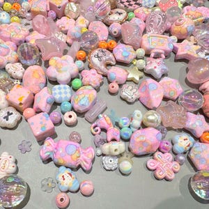 Pode incluir: Uma cole&ccedil;&atilde;o de contas rosa, brancas e iridescentes em v&aacute;rias formas, incluindo estrelas, flores e doces. Algumas contas t&ecirc;m detalhes e padr&otilde;es decorativos. As contas est&atilde;o espalhadas sobre uma superf&iacute;cie cinza claro.