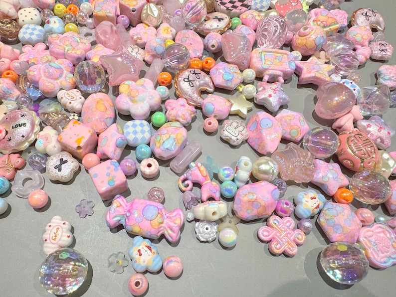 Pode incluir: Uma cole&ccedil;&atilde;o de contas rosa, brancas e iridescentes em v&aacute;rias formas, incluindo estrelas, flores e doces. Algumas contas t&ecirc;m detalhes e padr&otilde;es decorativos. As contas est&atilde;o espalhadas sobre uma superf&iacute;cie cinza claro.