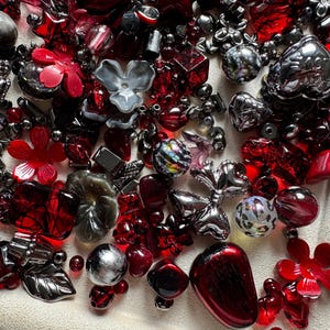 May include: A collection of assorted craft beads in various shapes and sizes, featuring a color palette of deep red, metallic silver, and charcoal gray. The assortment includes flower-shaped beads, heart-shaped charms, spheres, and faceted pieces.